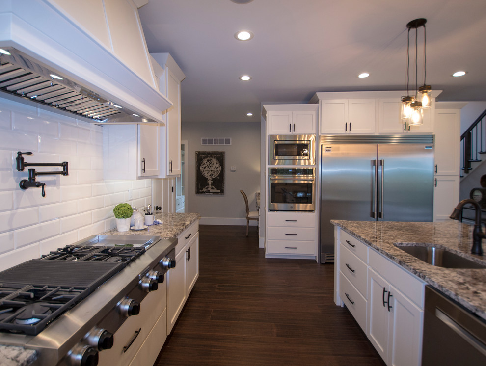 Design ideas for a large traditional u-shaped open plan kitchen in St Louis with shaker cabinets, white cabinets, granite benchtops, white splashback, subway tile splashback, stainless steel appliances, dark hardwood floors, with island, brown floor, beige benchtop and a farmhouse sink.