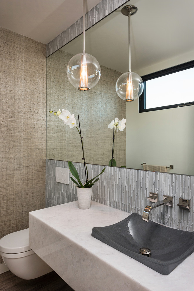 This is an example of a contemporary powder room in San Diego with a wall-mount toilet, gray tile, brown walls, a vessel sink, marble benchtops, brown floor, white benchtops, matchstick tile and dark hardwood floors.
