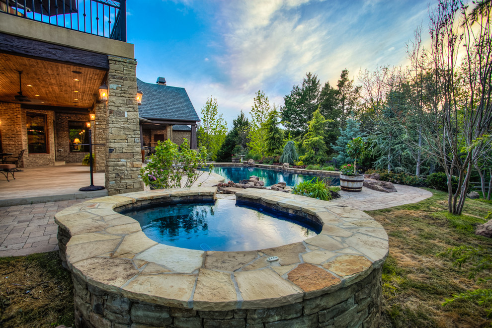 Photo of a large country backyard custom-shaped pool in Oklahoma City with a hot tub and natural stone pavers.