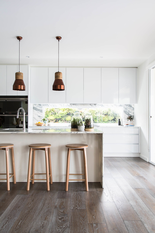 Design ideas for a scandinavian galley kitchen in Melbourne with flat-panel cabinets, white cabinets, window splashback, dark hardwood floors, with island, brown floor, marble benchtops, black appliances and white benchtop.