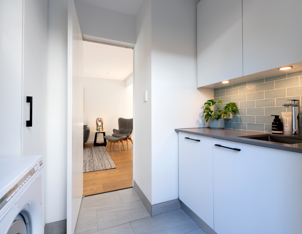 This is an example of a small contemporary galley dedicated laundry room in Melbourne with an undermount sink, flat-panel cabinets, white cabinets, quartz benchtops, subway tile splashback, white walls, porcelain floors, a stacked washer and dryer, grey floor and grey benchtop.