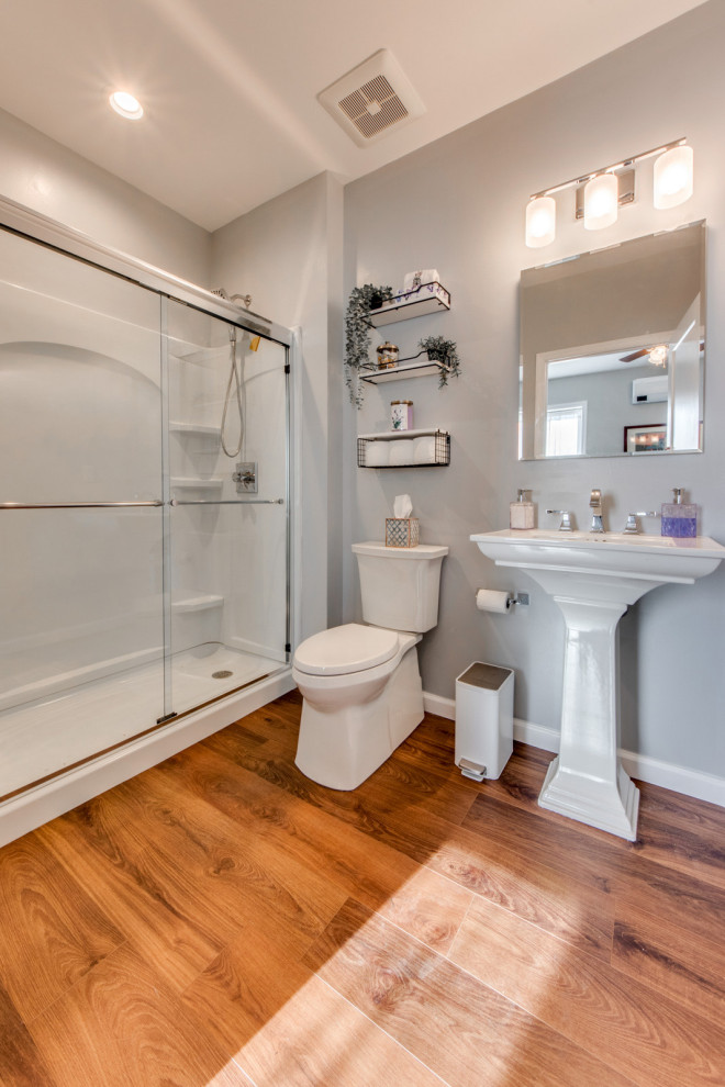 セントルイスにあるお手頃価格の中くらいなトランジショナルスタイルのおしゃれな浴室 (アルコーブ型シャワー、分離型トイレ、グレーの壁、ラミネートの床、ペデスタルシンク、茶色い床、引戸のシャワー、白い洗面カウンター、洗面台1つ) の写真