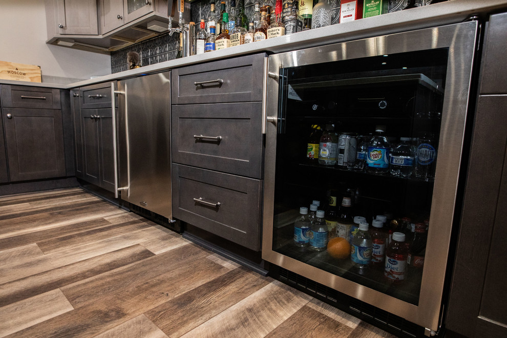 Photo of a small industrial u-shaped seated home bar in DC Metro with an undermount sink, shaker cabinets, medium wood cabinets, quartz benchtops, grey splashback, metal splashback, vinyl floors, brown floor and grey benchtop.