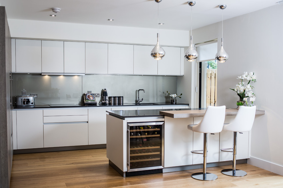 Inspiration for a contemporary l-shaped eat-in kitchen in Edinburgh with an undermount sink, flat-panel cabinets, white cabinets, grey splashback, glass sheet splashback, black appliances, medium hardwood floors, with island, brown floor and black benchtop.
