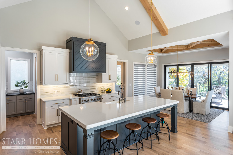 Photo of a beach style kitchen in Kansas City with shaker cabinets, white cabinets, grey splashback, stainless steel appliances, light hardwood floors, with island and black benchtop.