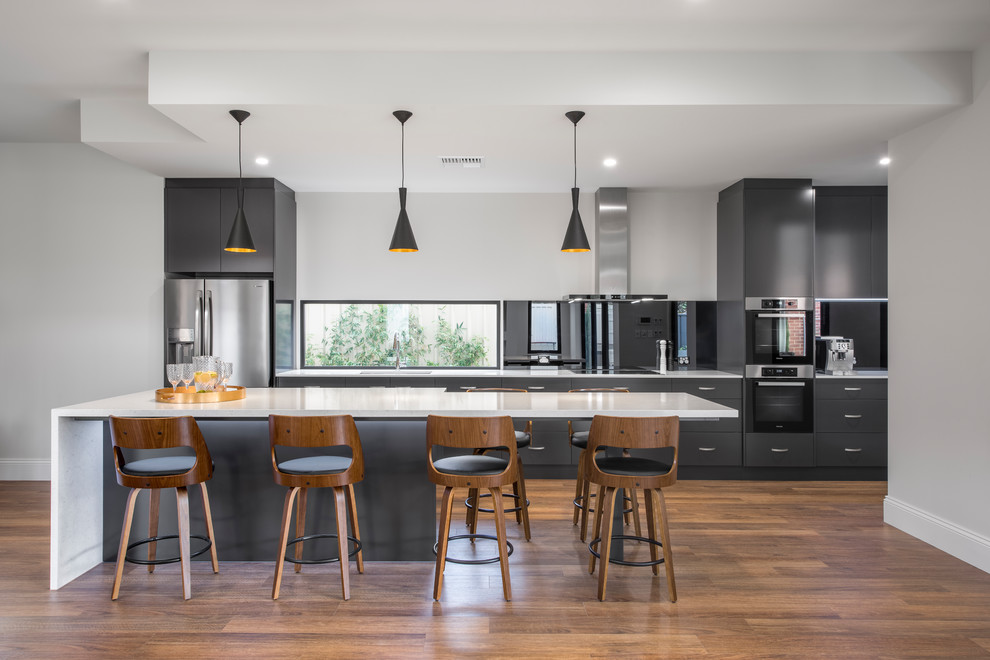 This is an example of a contemporary galley kitchen in Adelaide with flat-panel cabinets, grey cabinets, stainless steel appliances, medium hardwood floors, with island, brown floor and white benchtop.