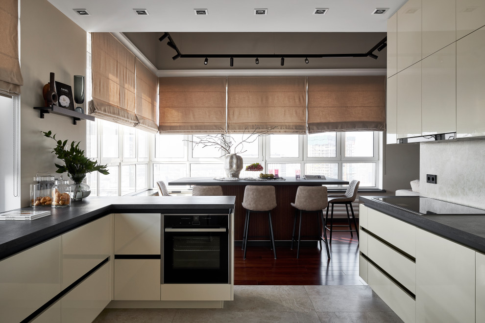 На фото: глянцевая кухня в современном стиле с бежевыми фасадами, серым фартуком, черной техникой, полом из керамогранита, островом, серым полом и черной столешницей с