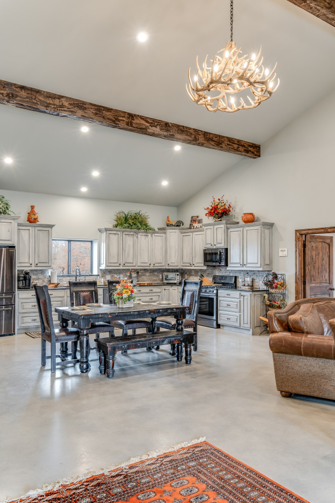 This is an example of a mid-sized country l-shaped eat-in kitchen in Austin with a drop-in sink, raised-panel cabinets, grey cabinets, granite benchtops, grey splashback, stone tile splashback, stainless steel appliances, concrete floors, grey floor, black benchtop and vaulted.