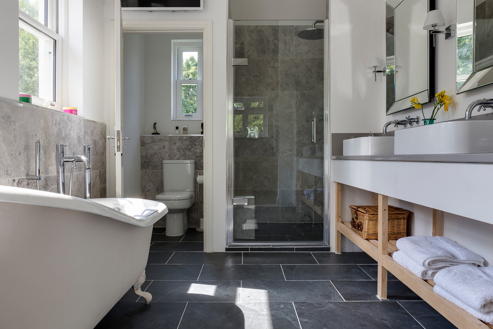 Inspiration for a large transitional kids bathroom in Wiltshire with a freestanding tub, a corner shower, black floor, a hinged shower door, open cabinets, light wood cabinets, a one-piece toilet, gray tile, porcelain tile, white walls, porcelain floors, a trough sink and stainless steel benchtops.