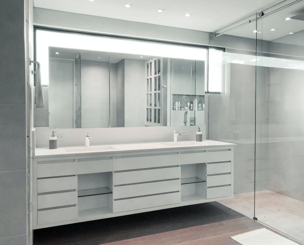 Inspiration for a medium sized modern ensuite bathroom with flat-panel cabinets, white cabinets, a double shower, a wall mounted toilet, grey walls, wood-effect flooring, a built-in sink, marble worktops, brown floors, a sliding door, white worktops, a wall niche, double sinks, a floating vanity unit and brick walls.