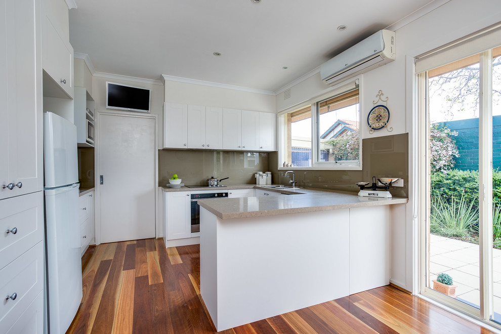 Small traditional u-shaped eat-in kitchen in Melbourne with a double-bowl sink, shaker cabinets, white cabinets, solid surface benchtops, beige splashback, glass sheet splashback, stainless steel appliances and dark hardwood floors.