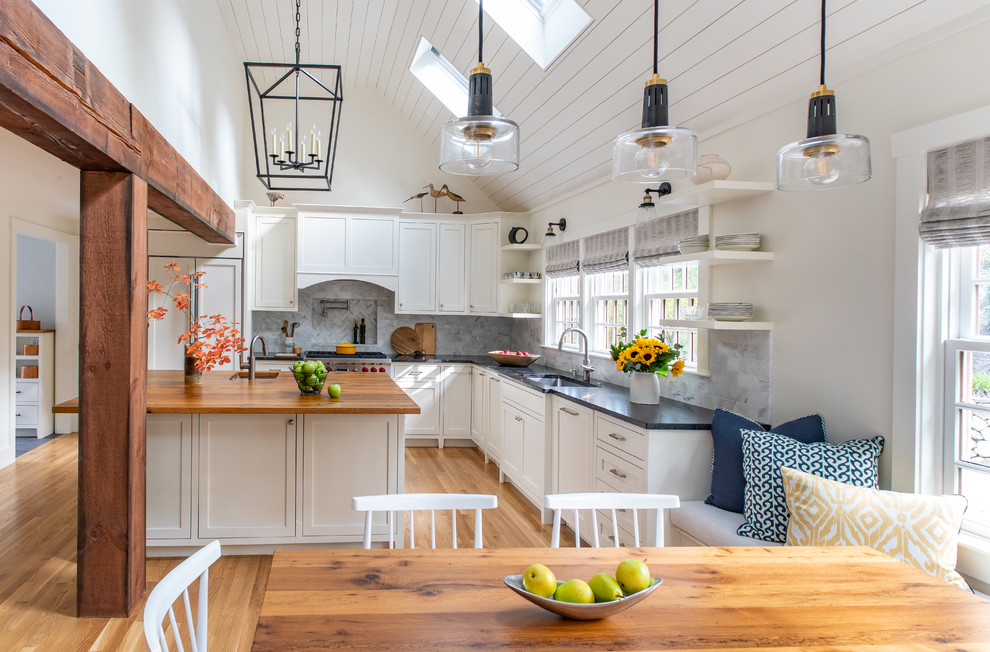Inspiration for a beach style l-shaped eat-in kitchen in Boston with shaker cabinets, white cabinets, with island, black benchtop, an undermount sink, grey splashback, light hardwood floors, beige floor, granite benchtops, marble splashback and panelled appliances.