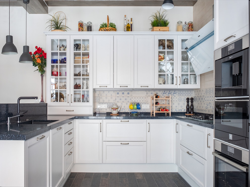This is an example of a large traditional u-shaped kitchen in Moscow with an undermount sink, white cabinets, quartz benchtops, ceramic splashback, black appliances, a peninsula, brown floor, black benchtop, glass-front cabinets, multi-coloured splashback and dark hardwood floors.