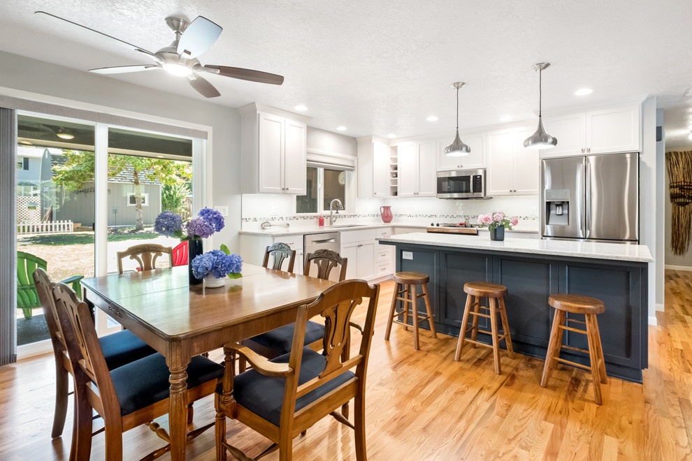 Example of a large transitional l-shaped medium tone wood floor and brown floor eat-in kitchen design with a drop-in sink, recessed-panel cabinets, white cabinets, quartzite countertops, white backsplash, ceramic backsplash, stainless steel appliances, an island and white countertops