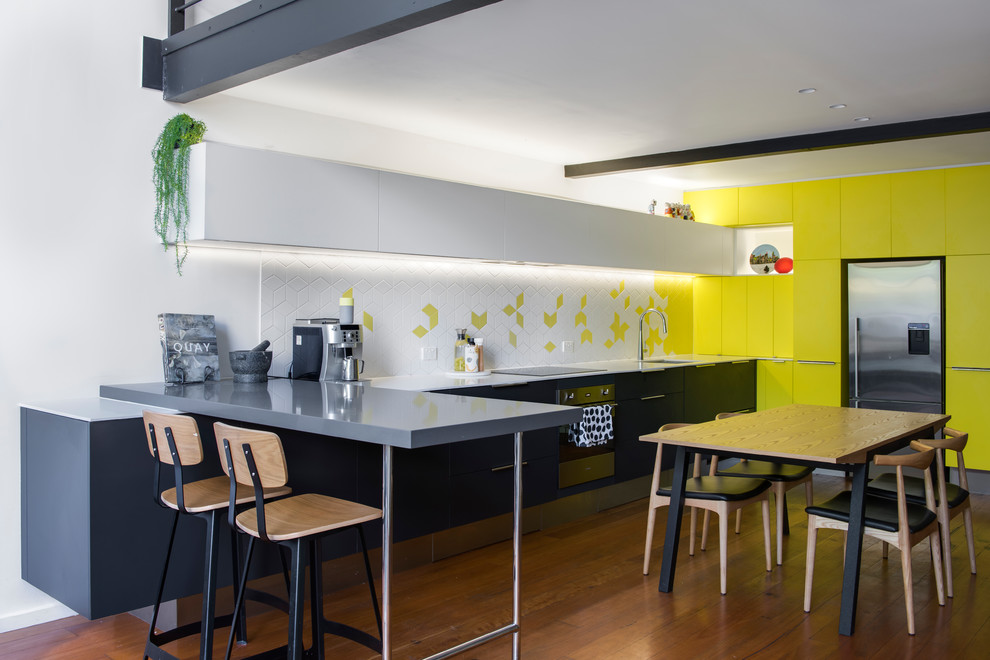 Small contemporary l-shaped open plan kitchen in Sydney with flat-panel cabinets, solid surface benchtops, white splashback, ceramic splashback, stainless steel appliances, medium hardwood floors, brown floor, an undermount sink, yellow cabinets and a peninsula.