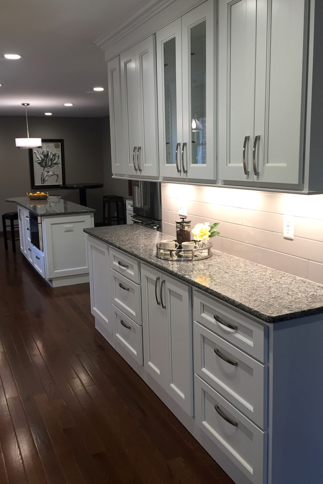 Transitional galley dark wood floor kitchen photo with an undermount sink, flat-panel cabinets, white cabinets, granite countertops, white backsplash, porcelain backsplash, stainless steel appliances and an island