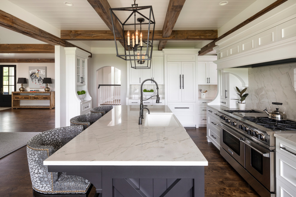 Photo of a farmhouse l-shaped kitchen in Minneapolis with a belfast sink, recessed-panel cabinets, white cabinets, white splashback, stone slab splashback, stainless steel appliances, dark hardwood flooring, an island, brown floors, white worktops, exposed beams and a timber clad ceiling.