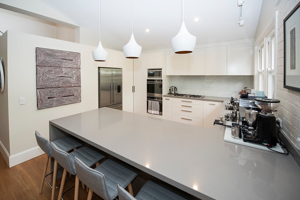 Photo of a mid-sized contemporary u-shaped eat-in kitchen in Sydney with an undermount sink, flat-panel cabinets, white cabinets, quartz benchtops, white splashback, porcelain splashback, stainless steel appliances, medium hardwood floors, a peninsula, grey benchtop and vaulted.