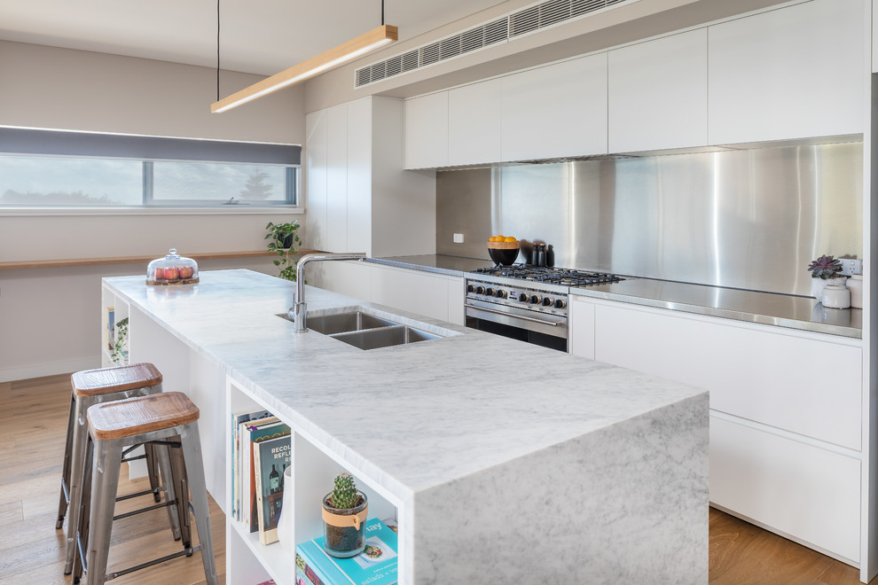 Inspiration for a contemporary galley open plan kitchen in Perth with a double-bowl sink, flat-panel cabinets, white cabinets, stainless steel benchtops, metallic splashback, stainless steel appliances, light hardwood floors, with island and grey benchtop.