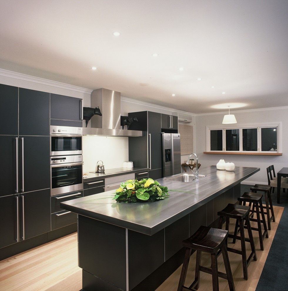 Inspiration for a contemporary single-wall eat-in kitchen in Wellington with a single-bowl sink, grey cabinets, stainless steel benchtops, white splashback, glass sheet splashback, stainless steel appliances, light hardwood floors and with island.
