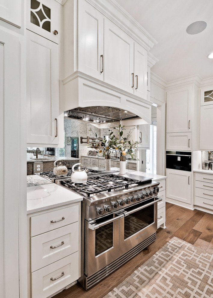 This is an example of a large transitional eat-in kitchen in Other with recessed-panel cabinets, white cabinets, quartzite benchtops, mirror splashback, stainless steel appliances, with island, white benchtop and light hardwood floors.
