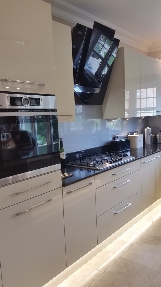 This is an example of a contemporary open plan kitchen in Gloucestershire with flat-panel cabinets, white cabinets, quartzite benchtops, metallic splashback, glass sheet splashback and limestone floors.