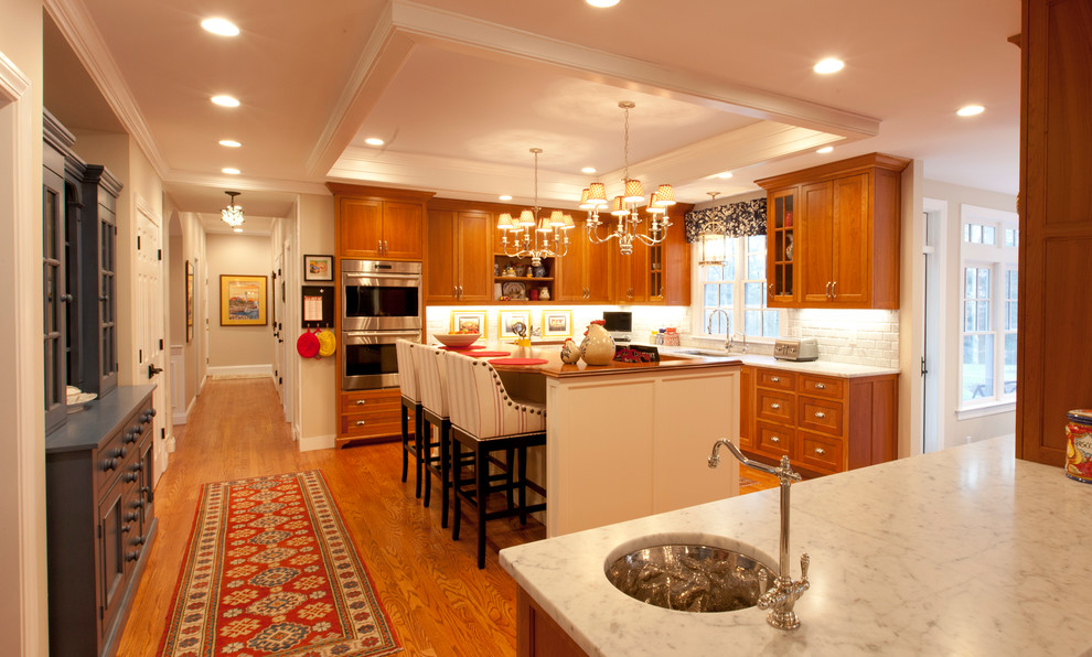 Inspiration for a large country l-shaped eat-in kitchen in Boston with an undermount sink, recessed-panel cabinets, orange cabinets, marble benchtops, white splashback, brick splashback, stainless steel appliances, medium hardwood floors, with island, brown floor and white benchtop.