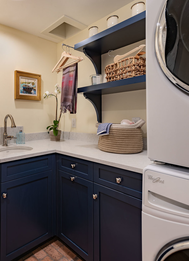 This is an example of a small traditional l-shaped dedicated laundry room in Boston with an undermount sink, recessed-panel cabinets, blue cabinets, quartz benchtops, yellow walls, brick floors, a stacked washer and dryer, red floor and grey benchtop.
