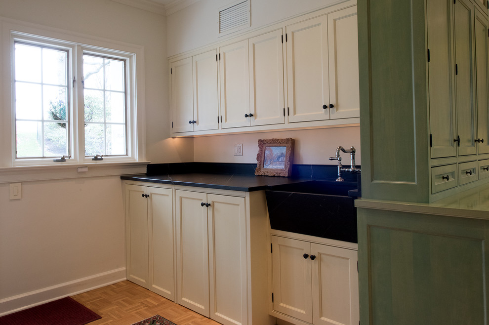 This is an example of a mid-sized traditional single-wall utility room in Philadelphia with a farmhouse sink, beaded inset cabinets, white cabinets, soapstone benchtops, white walls, light hardwood floors, a concealed washer and dryer, brown floor and black benchtop.