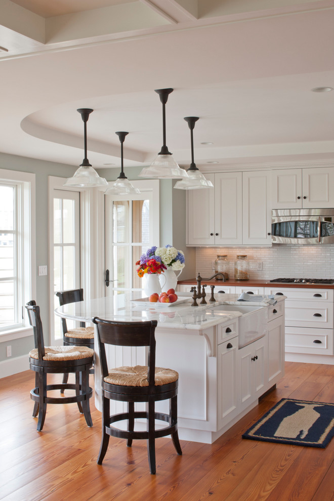 Inspiration for a mid-sized beach style l-shaped open plan kitchen in Boston with white cabinets, wood benchtops, white splashback, medium hardwood floors, a farmhouse sink, beaded inset cabinets, porcelain splashback, stainless steel appliances, with island and brown floor.