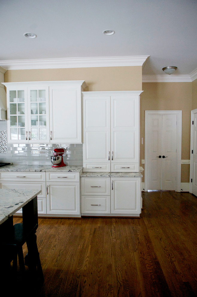 Inspiration for a mid-sized transitional l-shaped medium tone wood floor open concept kitchen remodel in Atlanta with a farmhouse sink, raised-panel cabinets, white cabinets, granite countertops, gray backsplash, glass tile backsplash, stainless steel appliances and an island