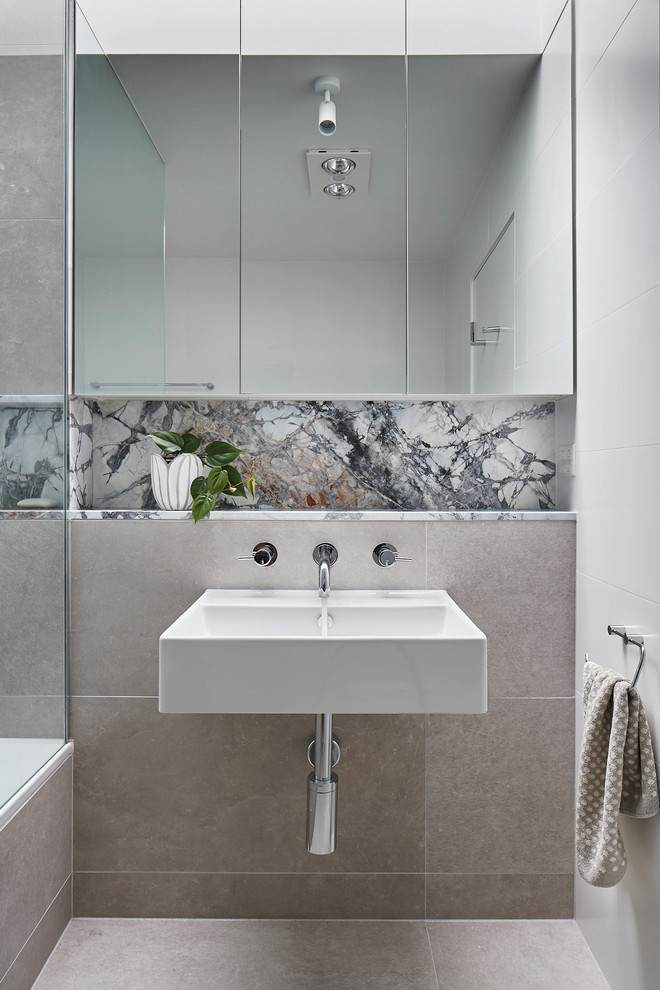 This is an example of a small modern bathroom in Melbourne with flat-panel cabinets, a drop-in tub, a shower/bathtub combo, a wall-mount toilet, beige tile, stone tile, white walls, porcelain floors, a wall-mount sink, marble benchtops, beige floor and white benchtops.