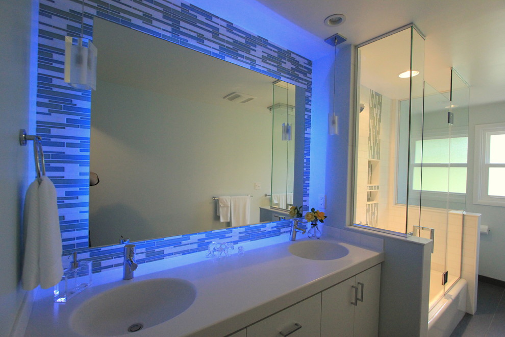 Design ideas for a mid-sized modern bathroom in Other with an integrated sink, flat-panel cabinets, white cabinets, a drop-in tub, a two-piece toilet, blue tile, mosaic tile, blue walls, ceramic floors, a shower/bathtub combo, grey floor, a hinged shower door, solid surface benchtops and white benchtops.