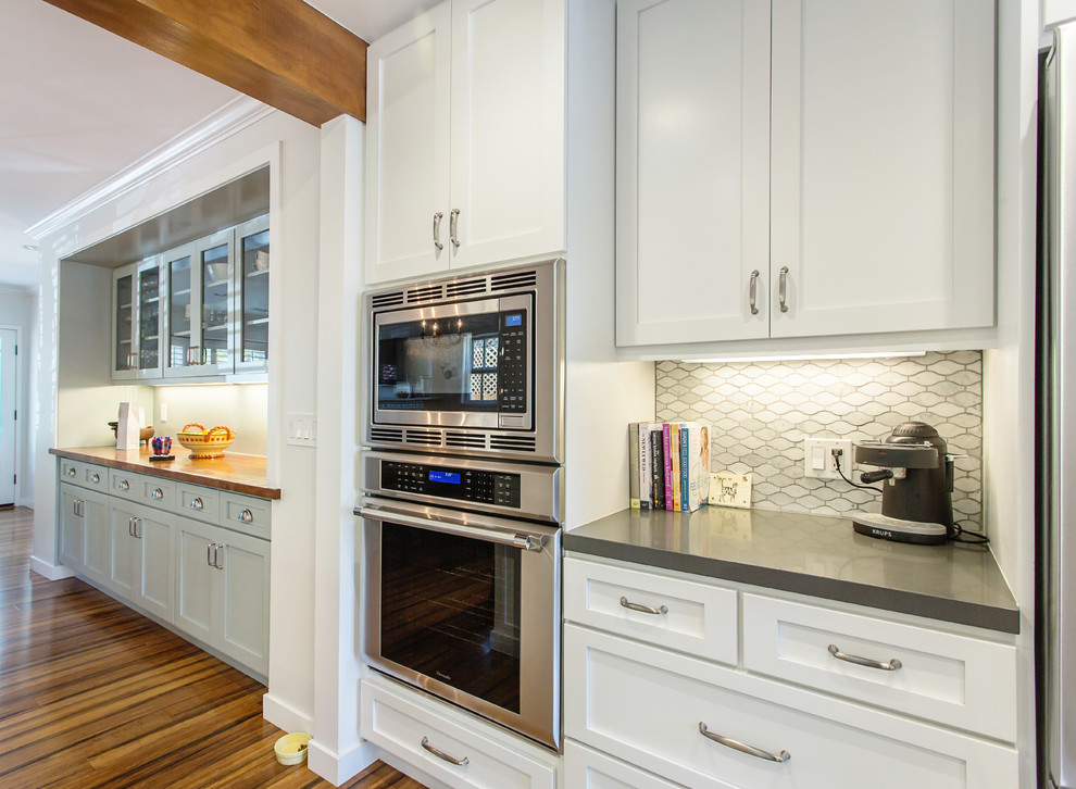 Photo of a mid-sized traditional u-shaped eat-in kitchen in Los Angeles with a farmhouse sink, shaker cabinets, white cabinets, quartz benchtops, grey splashback, stainless steel appliances, bamboo floors, porcelain splashback and a peninsula.