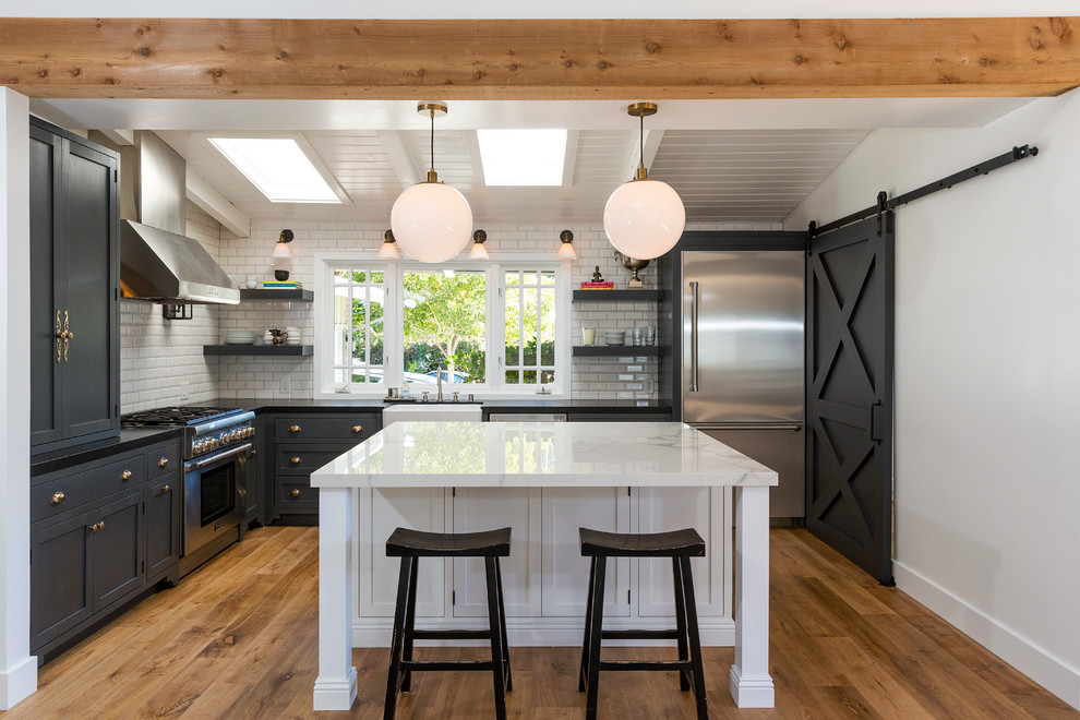 Photo of a beach style l-shaped kitchen in Los Angeles with a farmhouse sink, shaker cabinets, white splashback, subway tile splashback, stainless steel appliances, medium hardwood floors, with island and black cabinets.