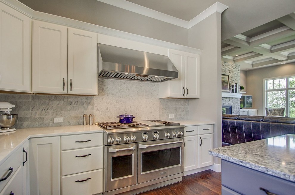 Example of a large transitional l-shaped medium tone wood floor kitchen design with a farmhouse sink, recessed-panel cabinets, white cabinets, stone tile backsplash, stainless steel appliances and an island