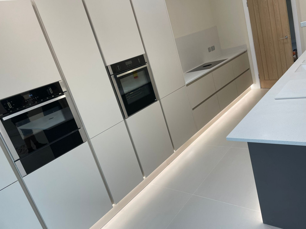 This is an example of a mid-sized contemporary single-wall open plan kitchen in Other with a drop-in sink, flat-panel cabinets, beige cabinets, marble benchtops, white splashback, marble splashback, black appliances, porcelain floors, with island, beige floor, white benchtop and vaulted.