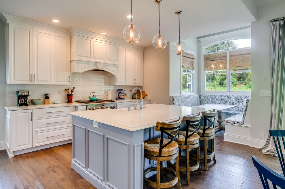 Design ideas for a beach style galley eat-in kitchen in Louisville with a farmhouse sink, raised-panel cabinets, white cabinets, white splashback, stainless steel appliances, medium hardwood floors, with island, brown floor and white benchtop.