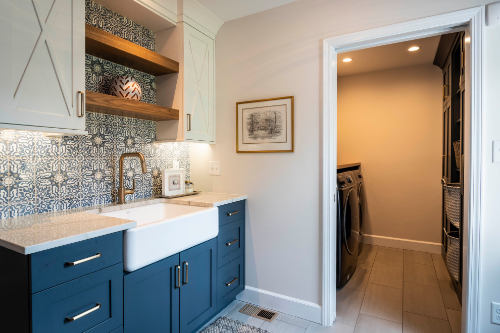 Design ideas for a large transitional utility room in Indianapolis with a farmhouse sink, shaker cabinets, blue cabinets, quartz benchtops, white walls, porcelain floors, a side-by-side washer and dryer, grey floor and white benchtop.