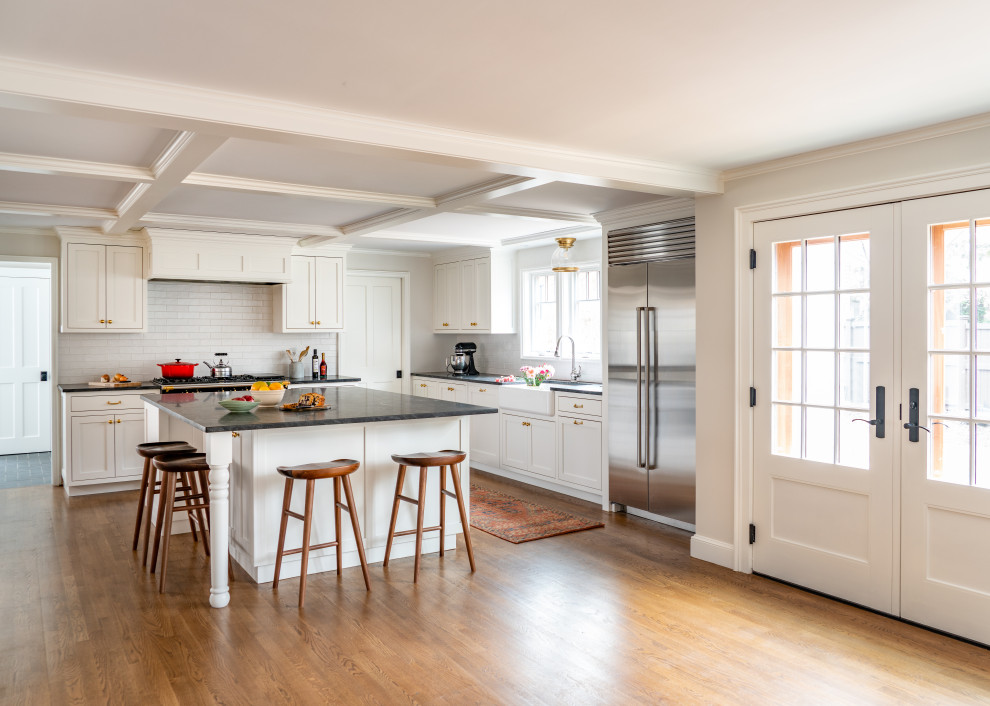 Mid-sized traditional l-shaped eat-in kitchen in Boston with a farmhouse sink, recessed-panel cabinets, white cabinets, white splashback, subway tile splashback, stainless steel appliances, medium hardwood floors, with island and black benchtop.