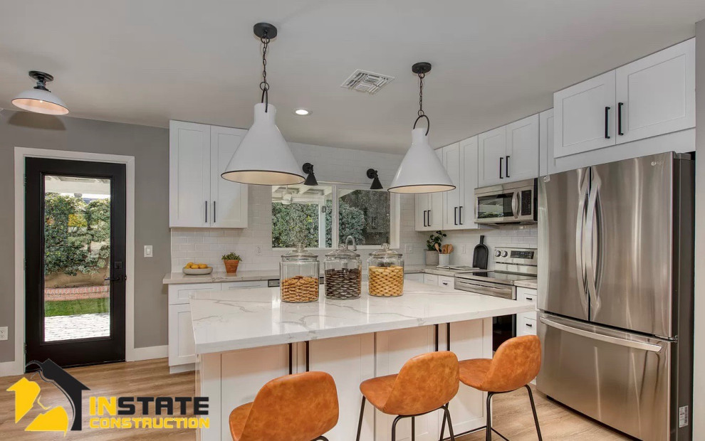 Large country l-shaped eat-in kitchen in Phoenix with a farmhouse sink, shaker cabinets, white cabinets, quartzite benchtops, white splashback, porcelain splashback, stainless steel appliances, light hardwood floors, with island, brown floor and white benchtop.