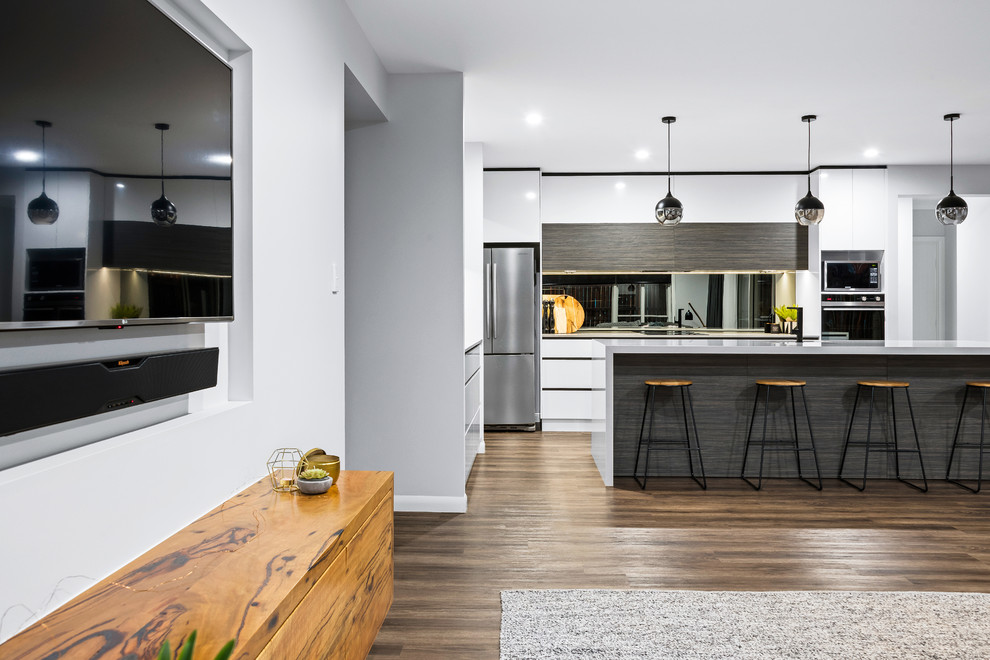 Mid-sized contemporary galley open plan kitchen in Other with an undermount sink, flat-panel cabinets, white cabinets, quartz benchtops, metallic splashback, mirror splashback, stainless steel appliances, laminate floors, with island, brown floor and white benchtop.