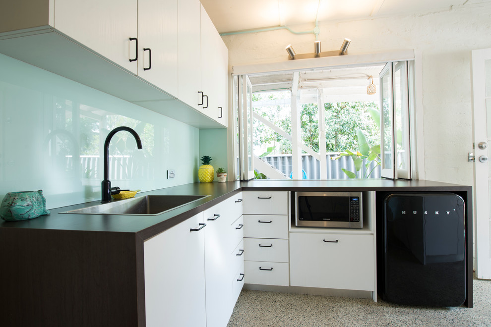 Photo of a small modern l-shaped eat-in kitchen in Perth with blue splashback, glass sheet splashback, a drop-in sink, flat-panel cabinets, white cabinets, no island, black appliances, bamboo floors and multi-coloured floor.