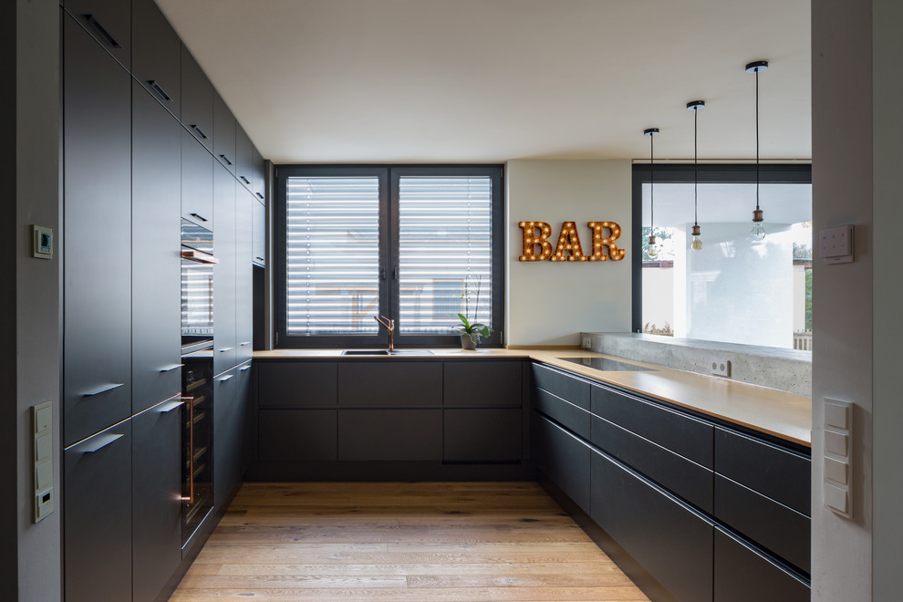 Photo of a mid-sized contemporary u-shaped open plan kitchen in Leipzig with an integrated sink, flat-panel cabinets, black cabinets, wood benchtops, black splashback, timber splashback, light hardwood floors, a peninsula, brown floor and black appliances.