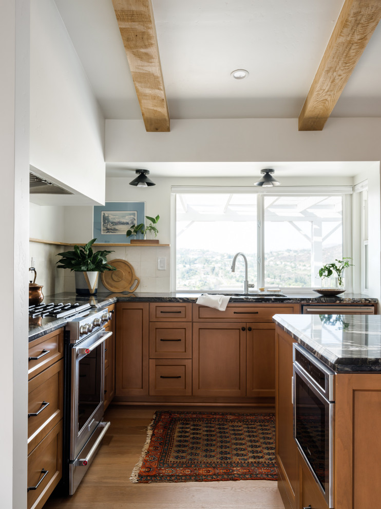 Design ideas for a mid-sized transitional l-shaped eat-in kitchen in San Diego with an undermount sink, raised-panel cabinets, light wood cabinets, granite benchtops, beige splashback, limestone splashback, stainless steel appliances, light hardwood floors, with island, beige floor, black benchtop and exposed beam.