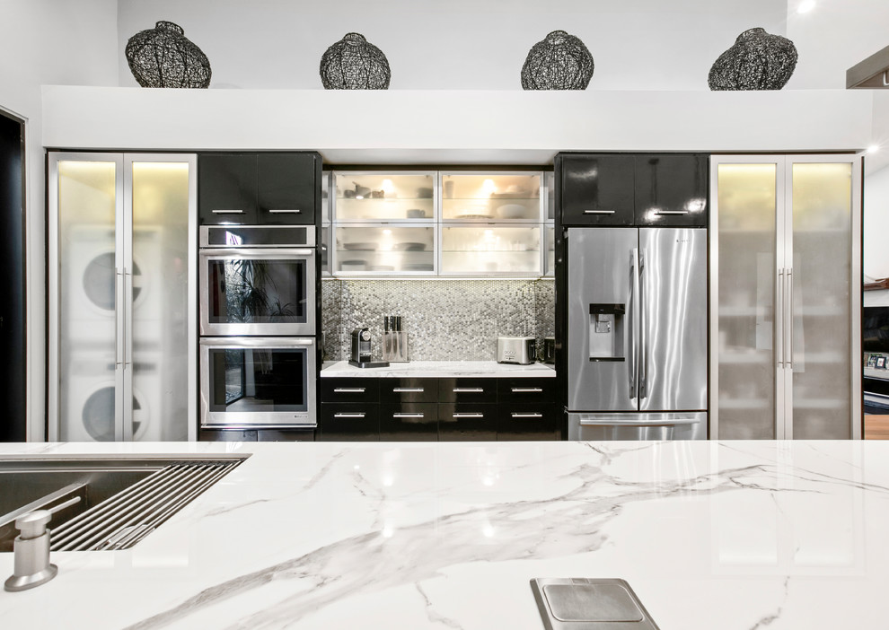 Photo of a large modern kitchen in Los Angeles with flat-panel cabinets, black cabinets, metallic splashback, metal splashback, stainless steel appliances, light hardwood floors, grey floor and white benchtop.