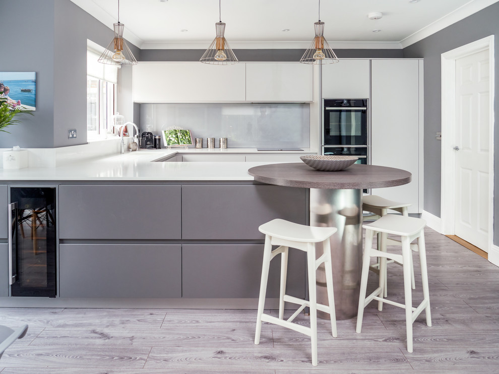 Design ideas for a contemporary u-shaped kitchen in Berkshire with a single-bowl sink, flat-panel cabinets, white cabinets, grey splashback, glass sheet splashback, black appliances, light hardwood floors, a peninsula and white benchtop.