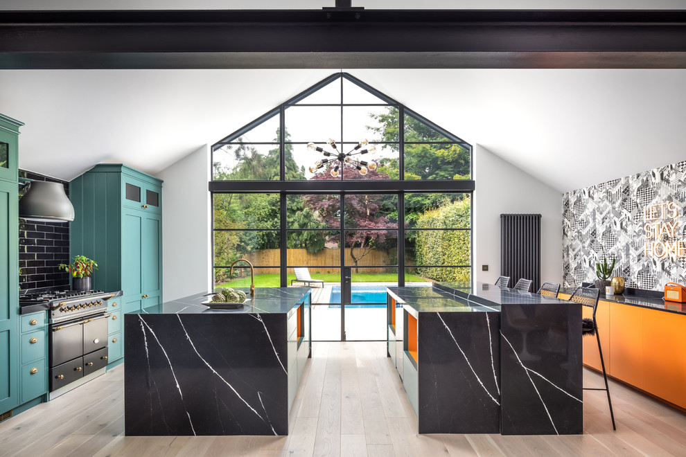 Photo of a contemporary galley open plan kitchen in Kent with multiple islands, shaker cabinets, blue cabinets, black splashback, black appliances, light hardwood floors, beige floor and black benchtop.