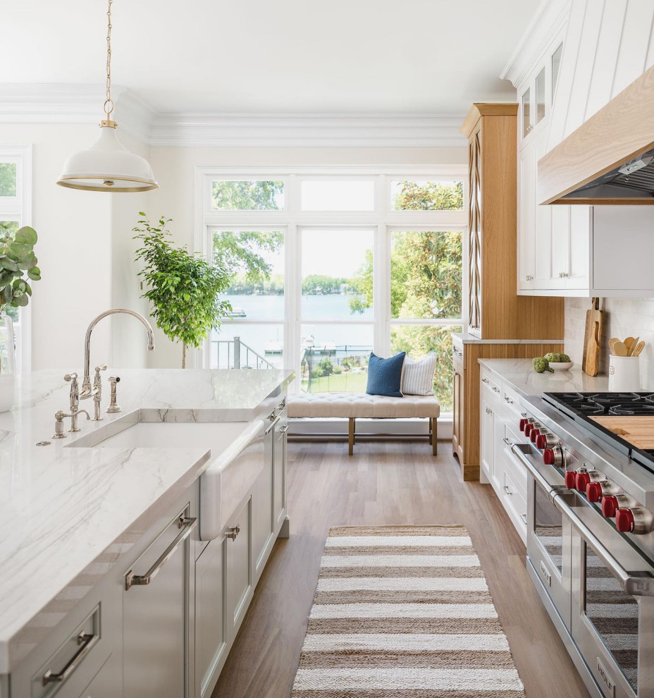 Inspiration for a large beach style l-shaped eat-in kitchen in Charlotte with a farmhouse sink, shaker cabinets, white cabinets, quartzite benchtops, grey splashback, marble splashback, stainless steel appliances, light hardwood floors, with island, beige floor and white benchtop.