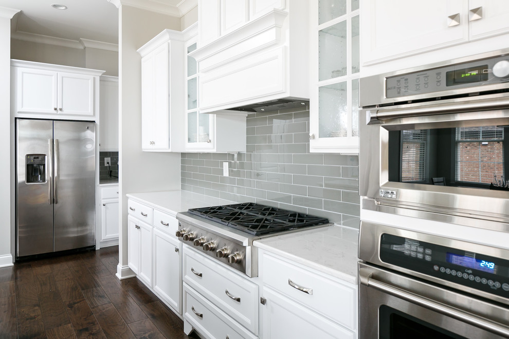 This is an example of a large transitional l-shaped eat-in kitchen in Louisville with an undermount sink, flat-panel cabinets, white cabinets, quartz benchtops, grey splashback, glass tile splashback, stainless steel appliances, dark hardwood floors and with island.